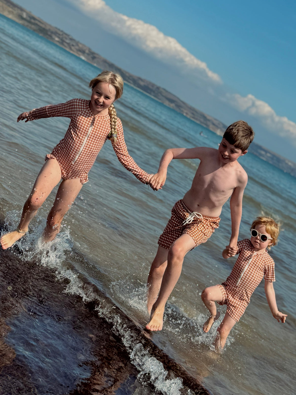 Three children running through the sea in GRECH & CO. Sienna Gingham swimwear, the youngest in round sunglasses