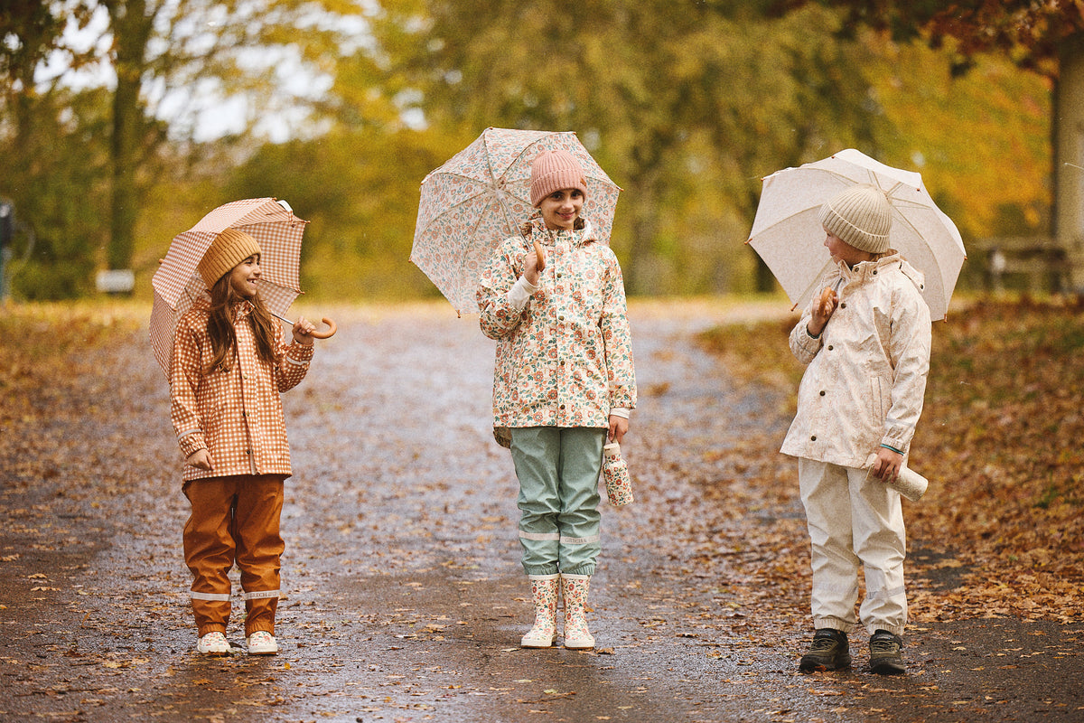 Three children on an autumn path in GRECH & CO. rainwear and knit beanies, holding Sienna Gingham, In Bloom and Pegasus umbrellas